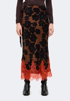 Modèle portant une jupe marron longueur cheville à imprimé floral noir, bordée de dentelle orange, et des bottines noires.