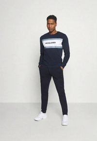 Marineblauwe crewneck trui met een horizontale wit- en blauwgestreepte afbeelding, logotekst, gecombineerd met marineblauwe joggerbroek en witte sneakers.