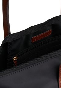 Približek notranjosti črne torbe, ki prikazuje žep z zadrgo, rjav usnjen trak in všit usnjen etiketo blagovne znamke znotraj.