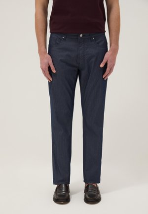 Hombre que lleva vaqueros azul oscuro de corte recto y mocasines marrón oscuro, de pie con las manos relajadas a los lados contra un fondo liso.