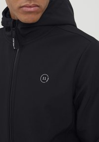 Crna zip jakna od mekog materijala, sa okruglim logotipom s brojem "11" i natpisom "Project" na lijevoj strani prsnog područja.