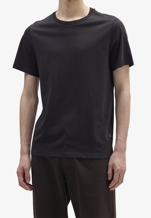 Persoon draagt een effen zwart T-shirt met korte mouwen en ronde hals en een donkere broek tegen een witte achtergrond.
