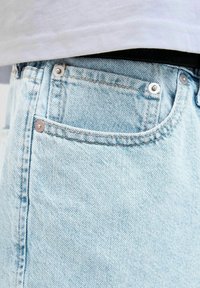 Jeans din denim de culoare albastru deschis cu o vedere detaliată a buzunarului din față, având rivete metalice argintii și detalii de cusătură portocalie.
