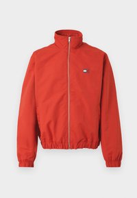 Κόκκινο zip-up μπουφάν από λείο ύφασμα, με ψηλό γιακά και ελαστικό στρίφωμα. Περιλαμβάνει ένα μικρό λογότυπο στην αριστερή πλευρά του στήθους.