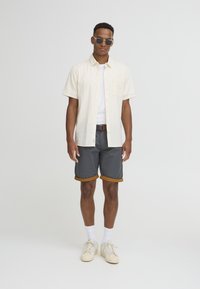 Light beige short-sleeve button-up shirt over a white T-shirt, paired with gray shorts with brown cuffs, and white sneakers. Sunglasses worn.