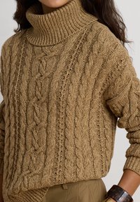 Femme portant un pull à col roulé beige en maille torsadée et un bracelet en écaille foncée à son poignet, avec de longs cheveux bruns visibles.