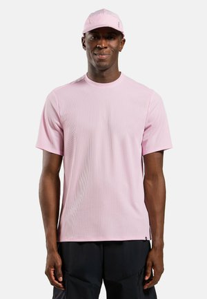 Homme portant une chemise à manches courtes rose clair et une casquette rose assortie, debout devant un fond blanc uni, les mains détendues le long du corps.