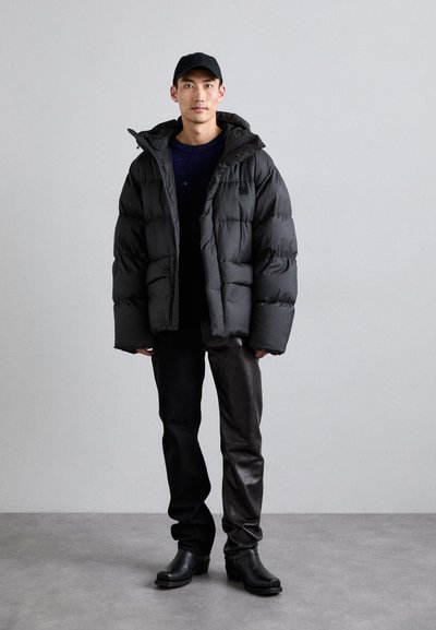 Chaqueta de plumas negra con capucha, que presenta un diseño acolchado. Lleva puesta sobre un suéter oscuro y se combina con pantalones de cuero negros y botas.