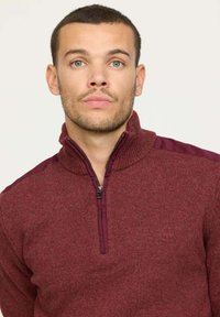 Sweat-shirt zippé bordeaux avec un col montant et une texture côtelée. Présente des accents d'épaule bordeaux foncé contrastants et une fermeture éclair argentée.