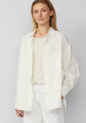 Witte, textuurrijke blouse met lange mouwen en knoopsluiting, gecombineerd met een lichtbeige top en witte broek, voorzien van een geweven riem.