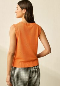 Top senza maniche lavorato a maglia arancione con scollo e orlo a costine. Abbinato a pantaloni a quadri bianco e nero. Tessuto liscio, vestibilità casual.