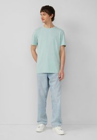 Mintgroene T-shirt met korte mouwen en ronde hals, gecombineerd met lichtblauwe straight-leg jeans en witte sneakers, staande tegen een neutrale achtergrond.