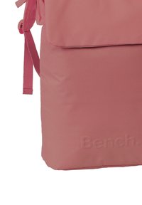 Sac à dos en tissu rose avec un rabat supérieur, des bretelles réglables et un logo "Bench" en relief sur le côté. Texture lisse, design minimaliste.