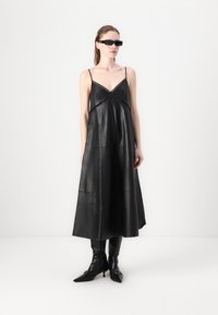 Schwarzes Lederkleid mit dünnen Trägern, V-Ausschnitt und Auschnitt-Details. Model trägt knielange schwarze Stiefel und Sonnenbrille. Glatte Textur.