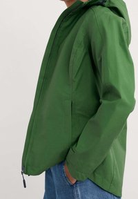 Zelená kapucí bunda s předním zipem, bočními kapsami a uvolněným střihem. Materiál má hladkou texturu a matný povrch.