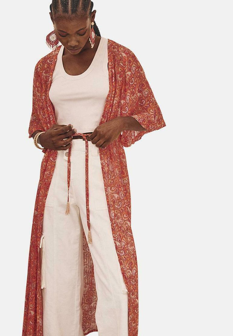 Kimono ligero de color rojo con estampado paisley, con mangas sueltas y atado en la cintura. Lleva puesto encima de una camiseta blanca ajustada y pantalones crema de pierna ancha.