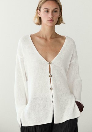 Donna che indossa un cardigan bianco lavorato a maglia ampio con grandi bottoni, scollo a V profondo e pantaloni neri, in piedi davanti a uno sfondo chiaro uniforme.