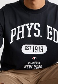 Černé bavlněné tričko s výrazným bílým textem "FYZ. VZD" a "ZALOŽENO 1919" s logem jako doplněk. Nosené s béžovými šortkami.