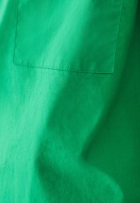 Kirkas vihreä kangas, jossa on sileä rakenne, varustettuna neliönmuotoisella läpällä ja siisteillä ompelu yksityiskohdilla.