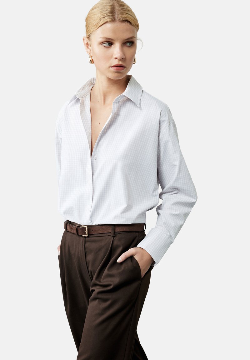 Camisa de cuadros blanca con cuello clásico, combinada con pantalones de tiro alto marrones y un cinturón marrón, que cuenta con bolsillos laterales.