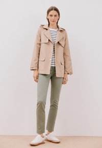 Beige lättviktig jacka med stora slag och framfickor, över en randig skjorta, kombinerad med olivgröna byxor och vita sneakers.