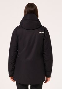 Veste noire imperméable avec un col haut et une capuche ajustable. Présente un logo au dos, un tissu lisse et un design ample.