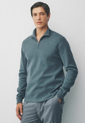 Suéter de media cremallera en mezcla de lana azul-verde, con puños y dobladillo acanalados. Combinado con pantalones de corte gris. Textura suave.