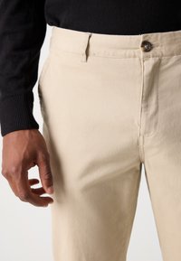 Beige cotton pants with a straight-leg fit, featuring a button closure, side pockets, and a soft texture. Worn with a black sweater.