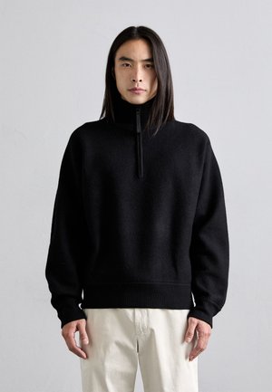 Black zip-neck sweater with a soft texture, dropped shoulders, and ribbed cuffs and hem, paired with off-white pants.
