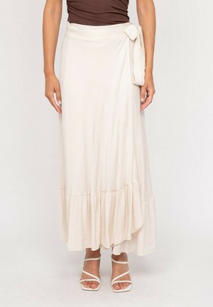 Femme portant une jupe portefeuille crème avec un lien sur le côté et un ourlet à volants, associée à des sandales à talons blanches à lanières et un haut marron.