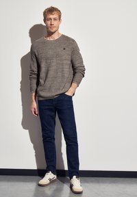 Muž v hnědém pleteném svetru, tmavě modrých džínech a béžových teniskách, stojící proti bílé zdi s jednou rukou v kapse.