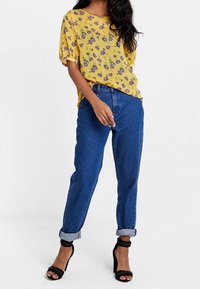 Blouse à motif floral jaune avec des manches courtes, associée à un jean en denim bleu à revers et des sandales à talons hauts noires.