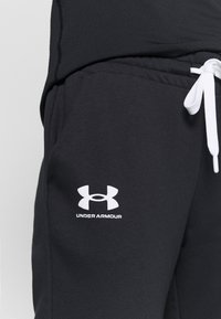 Svarta mjukisbyxor i mjukt material har en vit dragsko och en vit logotyp "Under Armour" nära vänster höft.