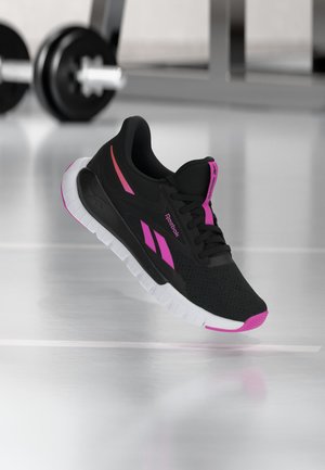 Zwart-roze Reebok sportschoen met witte zool zwevend boven een sportschoolvloer, met wazige sportgewichten op de achtergrond.