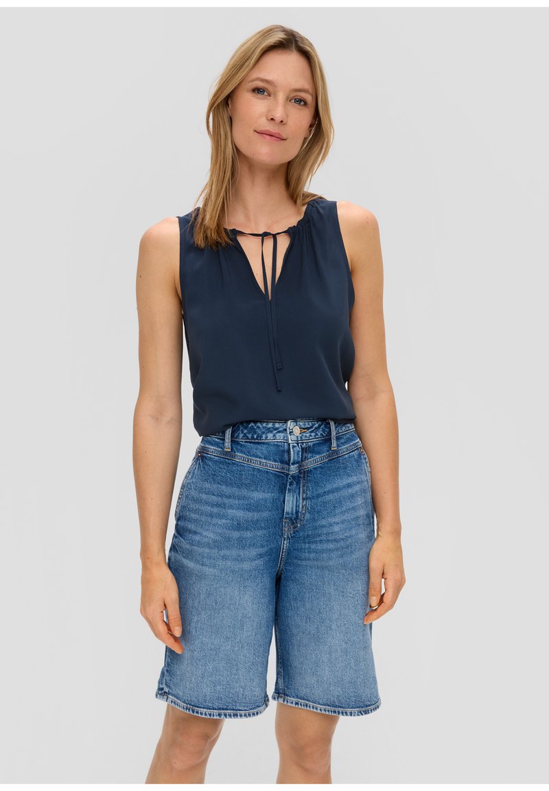 Blusa senza maniche di colore blu navy con scollo a V e dettaglio con fiocco, abbinata a pantaloni corti in denim blu a vita alta, con orlo sfilacciato.