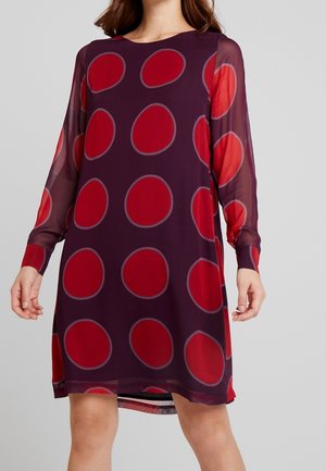 Langærmet knælang kjole i gennemsigtigt stof i mørklilla med store røde cirkelmønstre over det hele.