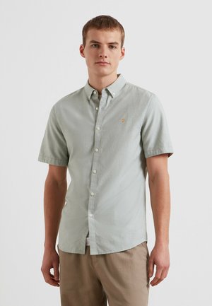 Giovane uomo che indossa una camicia leggera grigio chiaro a maniche corte e pantaloni beige, in piedi davanti a uno sfondo bianco uniforme.