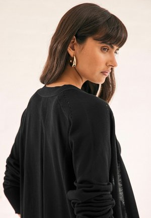 Schwarze Strickjacke mit gerippten Bündchen und nahtlosem Schulterdesign, kombiniert mit goldenen Tropfenohrringen. Sanfte Textur und lockere Passform.