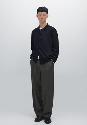 Schwarze, knöpfbare Strickjacke im lässigen Schnitt, über einem weißen Hemd getragen. Graue, plissierte Hose mit einem subtilen Muster, kombiniert mit schwarzen Schuhen.