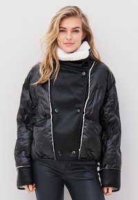 Fekete puffdzseki dupla gombolós kialakítással, kontrasztos fehér díszítéssel és magas gallérral. Puha, fényes felület nagy gombokkal.