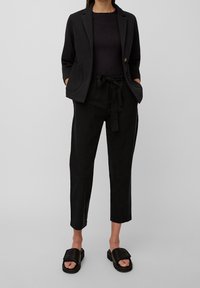 Blazer negro con bolsillos, combinado con una camiseta ajustada negra y pantalones sueltos negros con cordón. Sandalias deslizables negras completan el look.