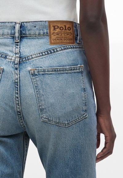 Φωτεινά μπλε τζιν από denim με πίσω τσέπες και καφέ δερμάτινη ετικέτα Polo Ralph Lauren, φορεμένα από άτομο με σκούρο δέρμα και λευκό πάνω μέρος.