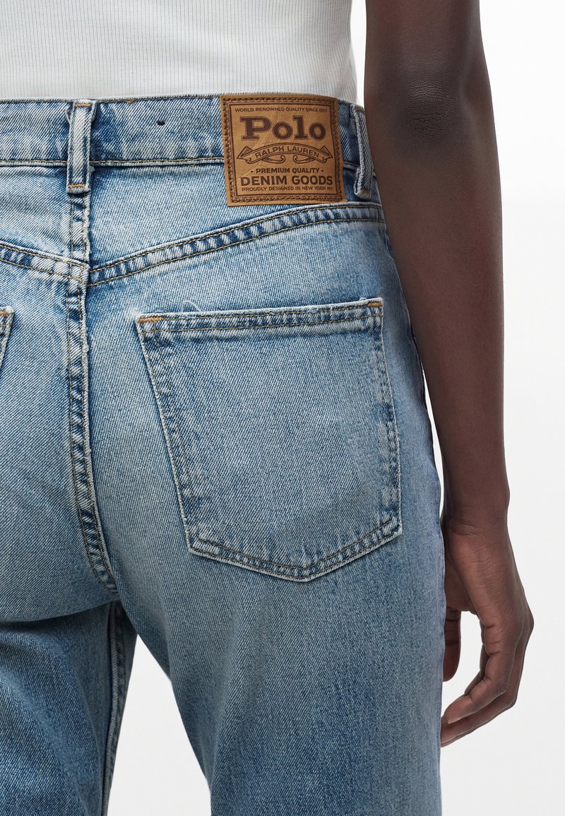 Jean en denim bleu clair avec poches arrière et un patch en cuir marron Polo Ralph Lauren porté par une personne à la peau foncée et un haut blanc.