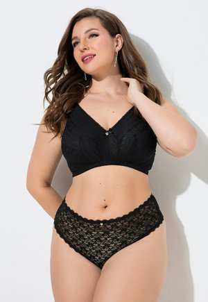 Kvinde med langt brunt hår iført sort blonde-bh og matchende højtaljede blonde-trusser, stående mod en hvid baggrund, smilende.