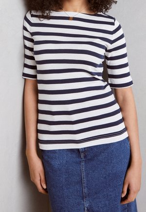Kvinde iført en marineblå og hvidstribet top med trekvartærmer og en blå denim nederdel, stående mod en lysegrå væg.