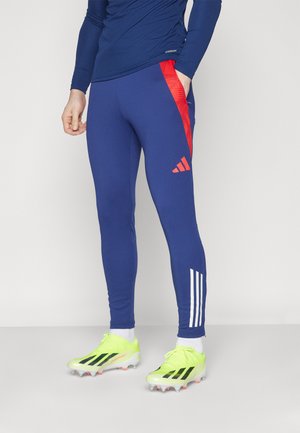 Blue athletic pants with red side accents, featuring a snug fit and subtle texture, paired with bright yellow soccer cleats.