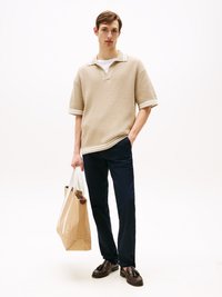 Ung mann iført en beige kortermet strikket skjorte, marineblå bukser, mørke loafers, holder en stor beige totebag, står mot en ensfarget hvit bakgrunn.