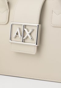 Beige taske med en glat tekstur og en fremtrædende sølvlogo-låse med "AX" initialer. Inkluderer kontraststing detaljer.