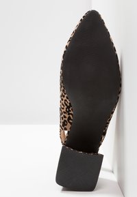 Chaussure à bout pointu avec empeigne imprimée léopard et semelle noire texturée ainsi qu’un talon bloc placée contre une surface d’angle blanche.