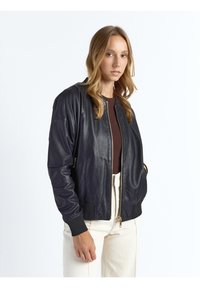 Zwarte leren bomberjack met een ritssluiting aan de voorkant, geribbelde manchetten, zijzakken en een gladde textuur, gedragen over een bruin shirt en lichte broek.